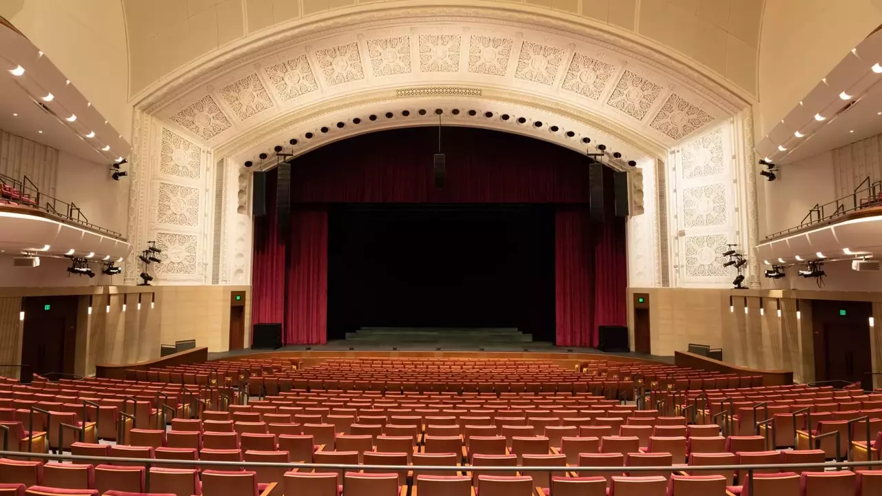 Empty auditorium with red seats facing a dark stage, framed by ornate architectural details and curtains.