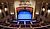 Theater interior featuring a blue stage, red curtains, and rows of empty seats, illuminated by overhead lights.
