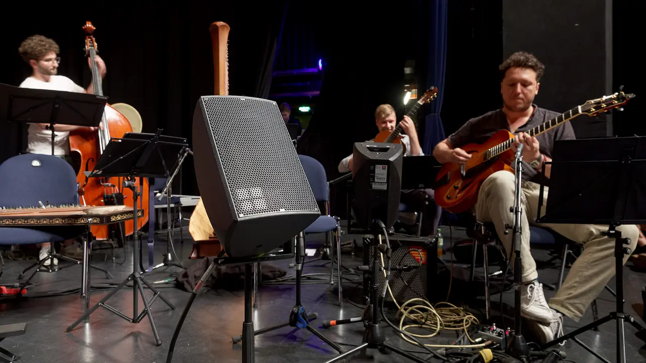Musiker, die auf der Bühne mit einem Kontrabass, einer Harfe und einer Gitarre auftreten, umgeben von Notenständern und Audioequipment.