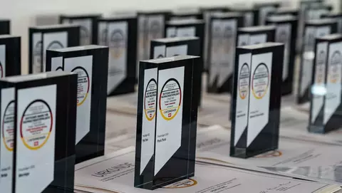 Multiple awards and certificates are displayed on a table, featuring clear glass trophies and printed documents with various names and details.