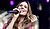 A woman with long, wavy hair sings into a microphone, wearing a pink outfit, with a joyful expression on her face.
