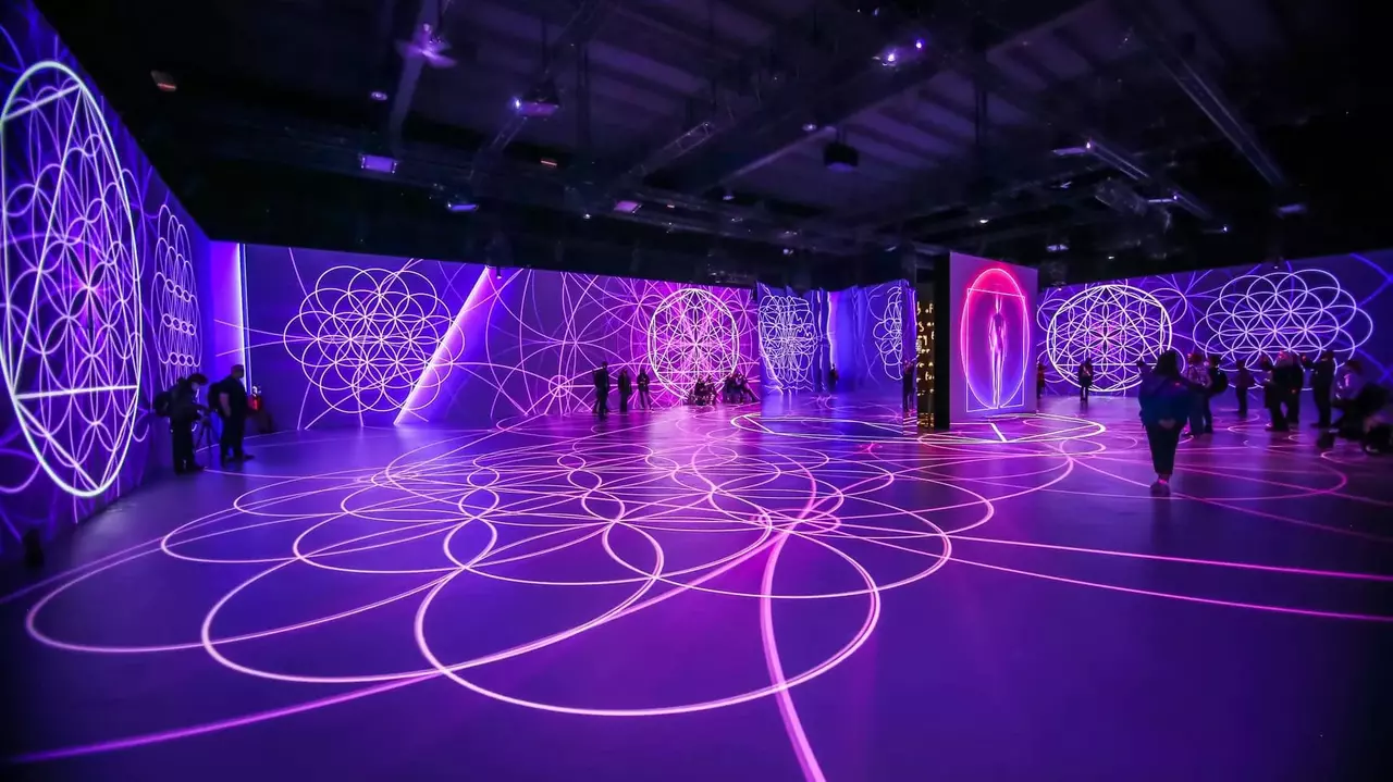 A large indoor space featuring vibrant purple and blue geometric light projections on the walls and floor, with people interacting throughout.