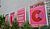 Four large pink posters titled "THE CAMPUS" display abstract designs and shapes, surrounded by greenery along a modern building facade.