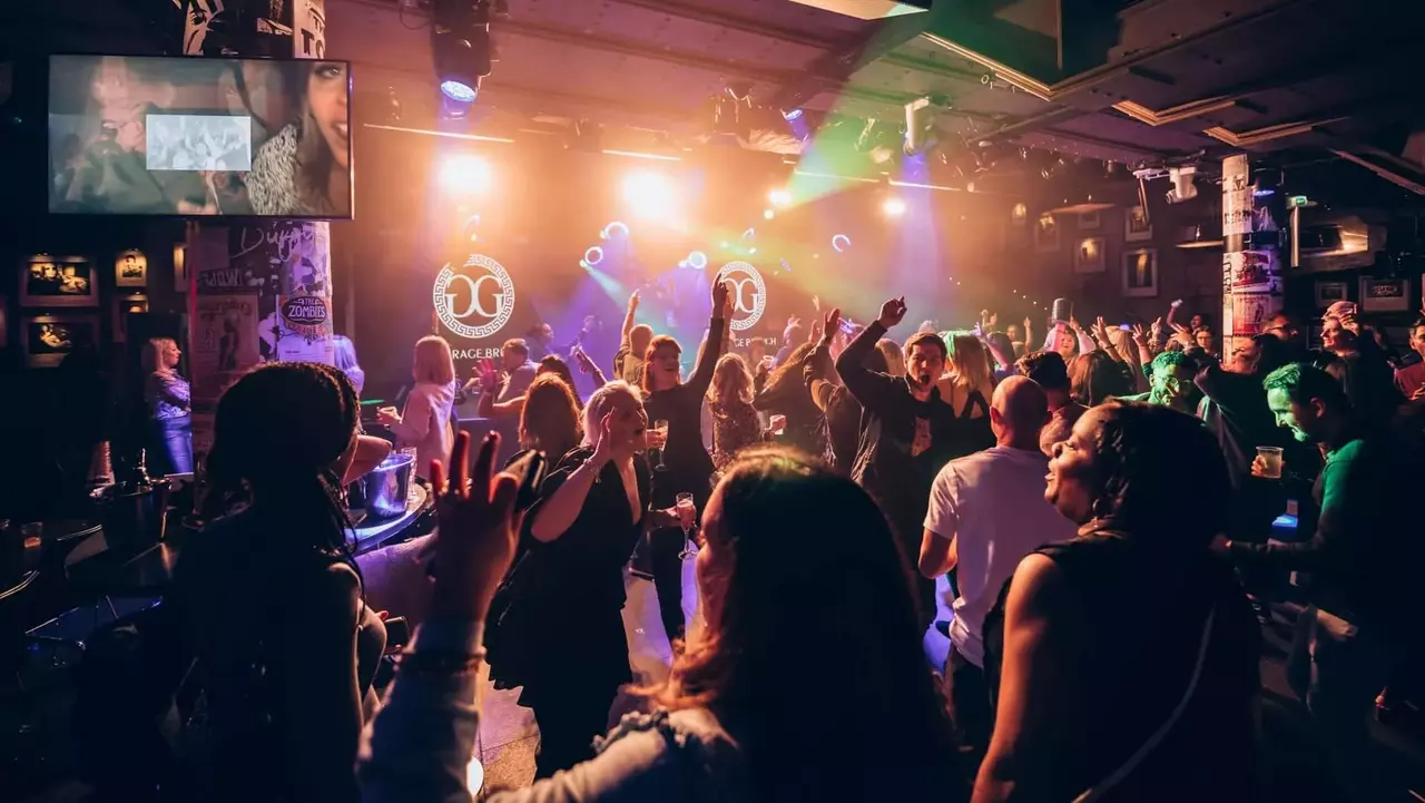 Crowd dancing and enjoying music at a lively venue, with colorful lights and a screen displaying visuals in the background.