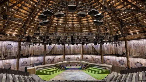 A large, circular auditorium with a wooden ceiling, featuring a stage at the center and rows of empty seats arranged around it.