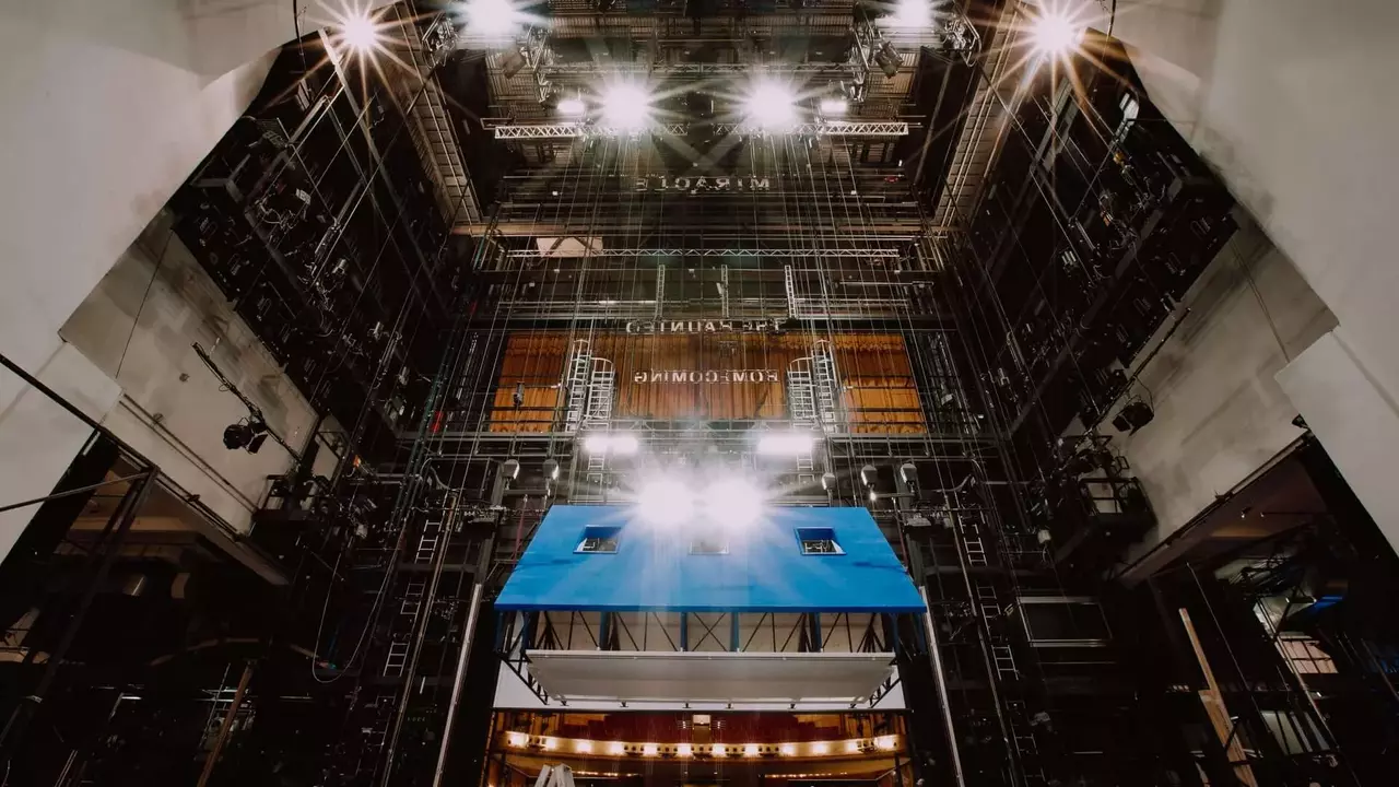 The interior of a theater stage, featuring overhead lights, rigging, and a blue backdrop, with a ladder visible in the foreground.