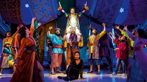 A vibrant musical performance featuring a diverse cast, with one performer elevated in the center, surrounded by others in colorful costumes.