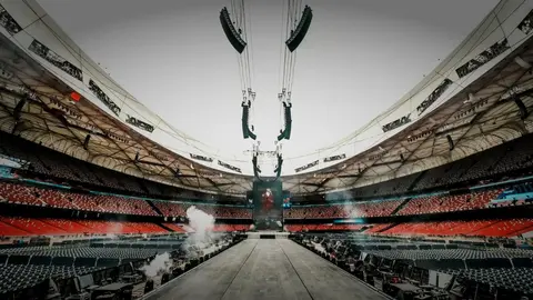 A wide view of a large stadium with empty seats, featuring a central stage and overhead sound equipment, set for an upcoming event.