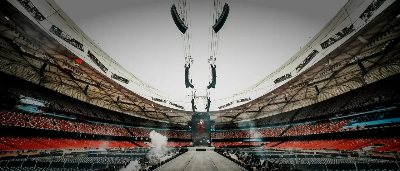 A wide view of a large stadium with empty seats, featuring a central stage and overhead sound equipment, set for an upcoming event.