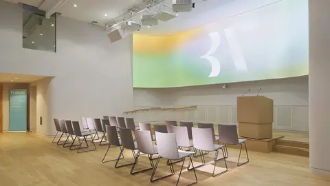 Modern auditorium with rows of chairs facing a stage, featuring a large, softly lit backdrop and audio equipment overhead.