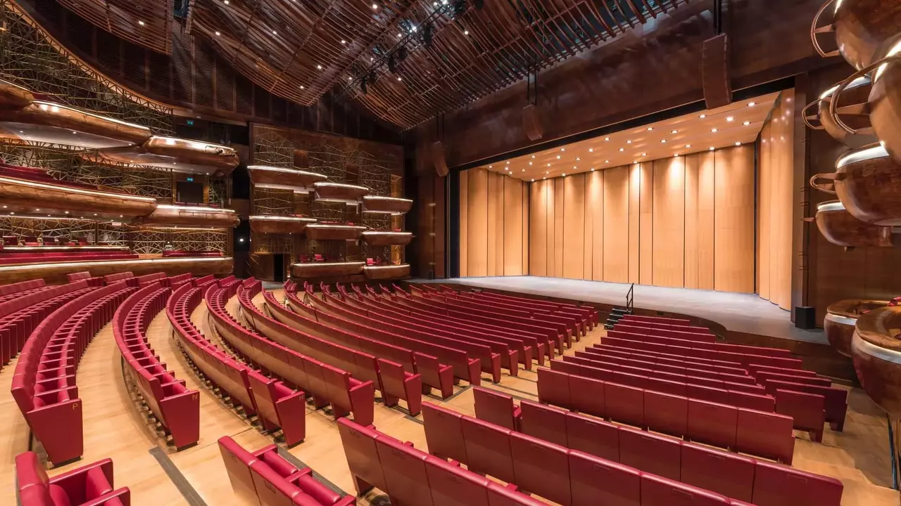 Modern auditorium with red seating arranged in a semi-circle, featuring a wooden stage and intricate architectural details.