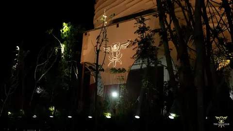 A modern building illuminated at night, featuring a stylized logo projected on its exterior, surrounded by trees and soft lighting.