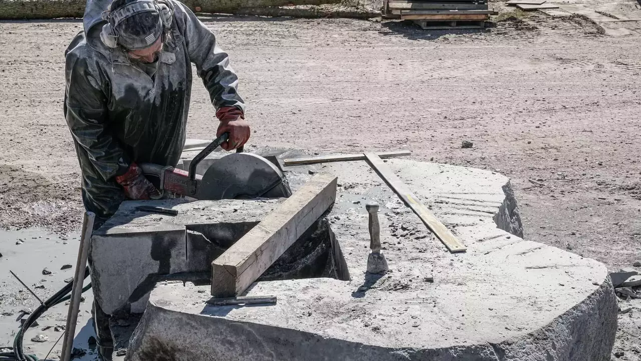 A sculptor works on a large stone block, using a tool to shape it, surrounded by tools and materials on the ground.