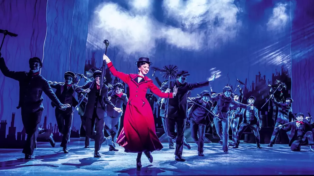 A performer in a red coat leads a group of dancers in a theatrical scene, set against a dramatic backdrop with a cloudy sky.