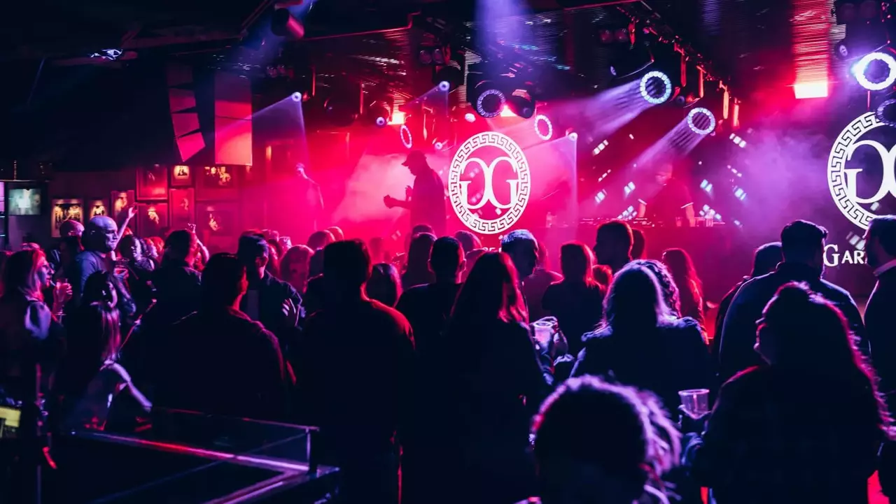 Crowd enjoying a live music performance at a nightclub, illuminated by colorful lights and featuring a prominent logo on stage.