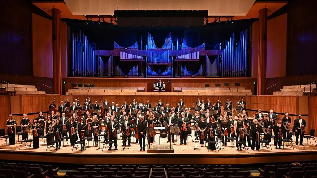 A large orchestra stands on stage, arranged in rows with various instruments, in a concert hall featuring a prominent organ in the background.