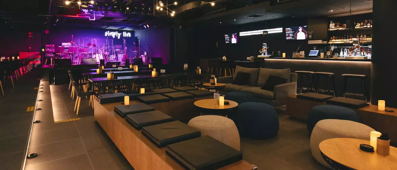 Modern lounge with seating areas, wooden tables, and a bar. A stage with musical instruments is visible in the background, illuminated by colorful lights.