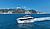 A luxury yacht navigates through clear blue waters near a rocky coastline with green hills in the background.