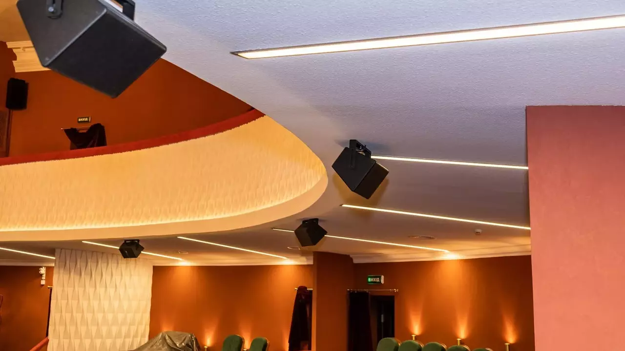 Deckenmontierte Lautsprecher und moderne Beleuchtungskörper in einem Theatersetting, mit einer geschwungenen architektonischen Gestaltung und grünen Sitzplätzen, die darunter sichtbar sind.