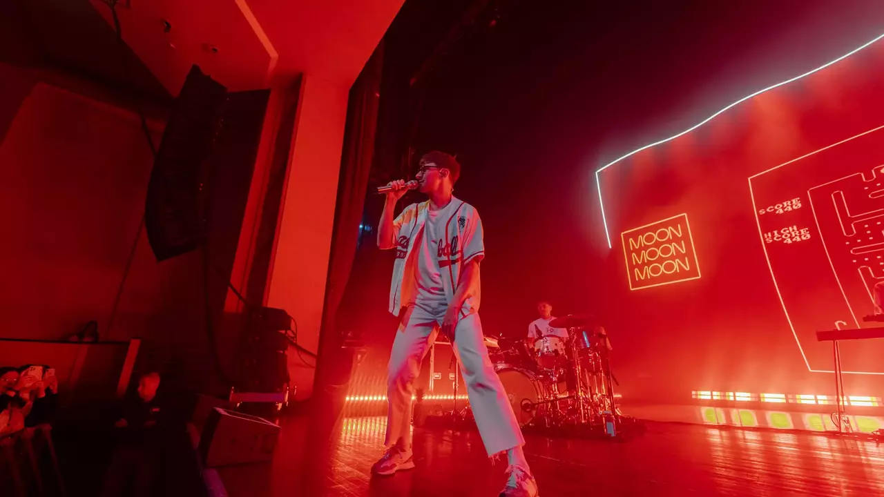 A performer sings into a microphone on stage, with a drummer in the background and vibrant red lighting illuminating the scene.
