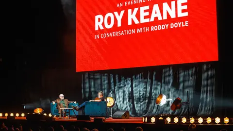 Roy Keane and Roddy Doyle seated on stage during a conversation event, with a large red screen displaying the event title behind them.