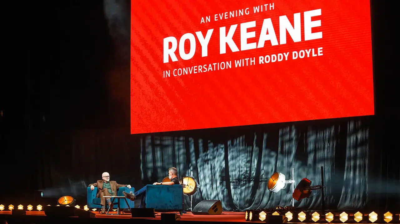 Roy Keane and Roddy Doyle seated on stage during a conversation event, with a large red screen displaying the event title behind them.