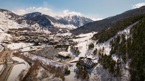 Snow-covered mountains surround a valley town, featuring ski slopes, buildings, and a parking area, under a clear blue sky.