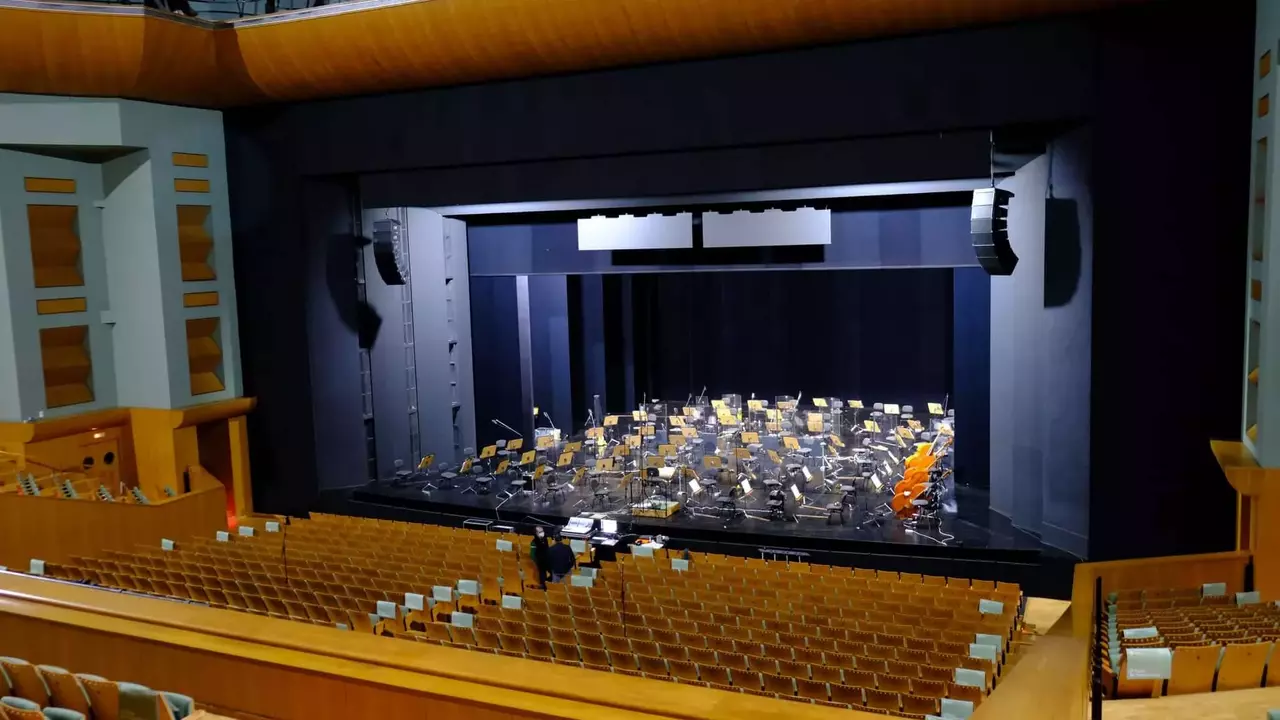 Empty concert hall with a stage set for an orchestra, featuring rows of chairs and music stands, illuminated by stage lights.