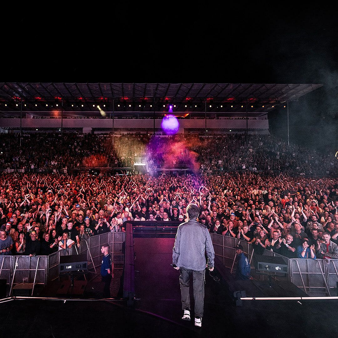 A performer stands on stage facing a large crowd at a concert, with vibrant lights illuminating the audience and venue.