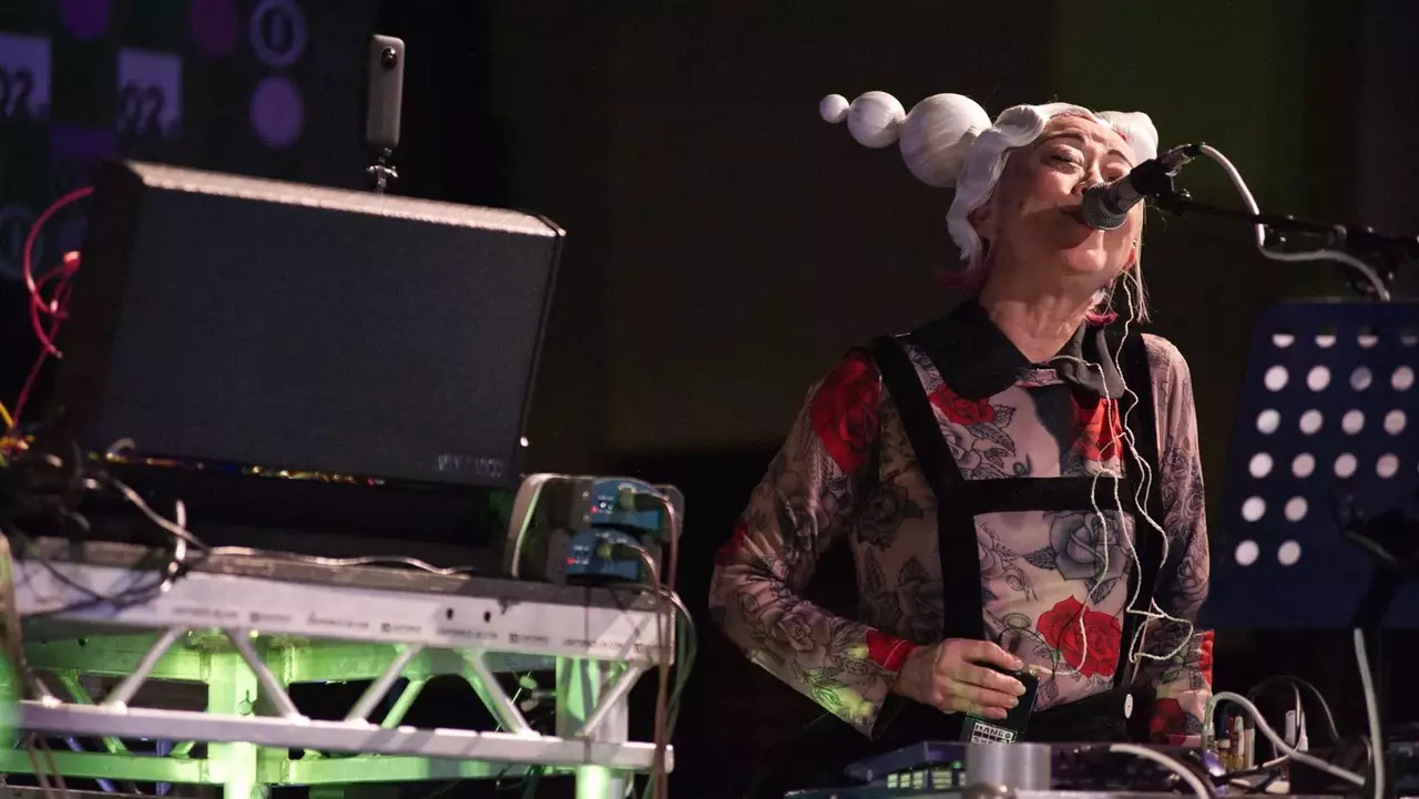 A performer with a unique hairstyle and floral outfit sings into a microphone while standing behind electronic equipment on stage.