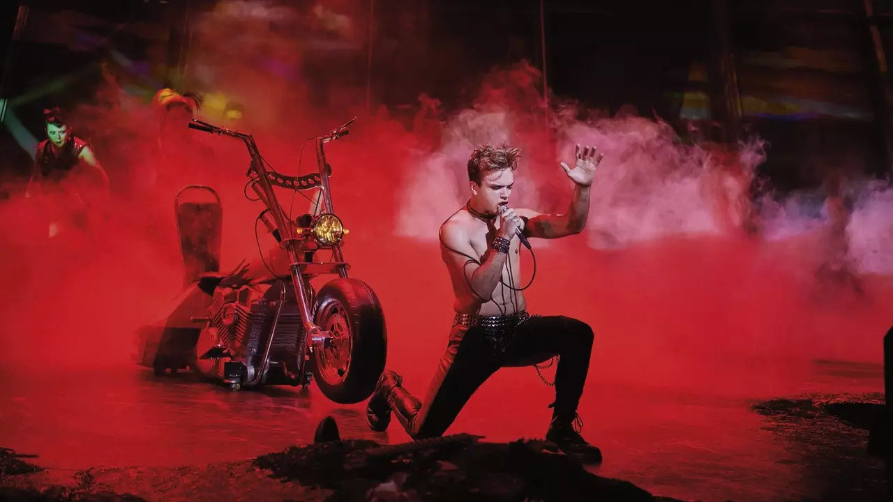 A performer kneels on stage, singing passionately into a microphone, with a motorcycle and smoke effects in the background.