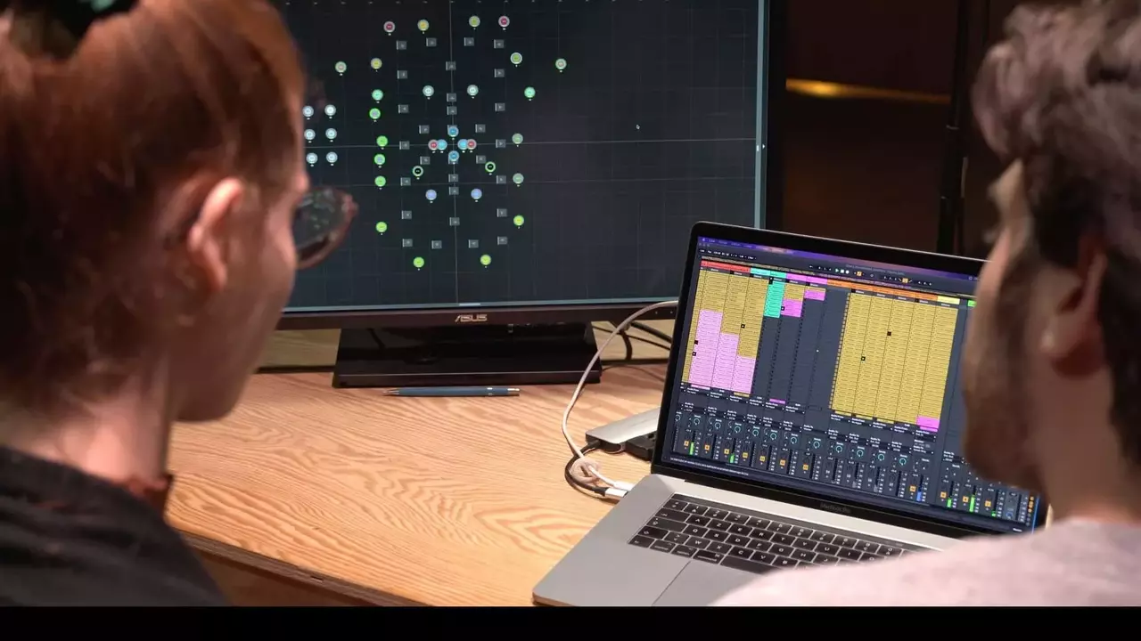 Two individuals are focused on a laptop displaying audio software, while a large monitor shows a visual representation of data points.