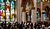 Crowd of people attending a service inside a church, with colorful stained glass windows illuminating the interior.