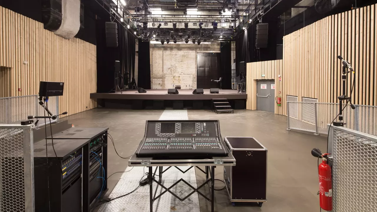 Empty performance venue with a sound mixing console in the foreground, wooden paneling on the walls, and a stage at the back.