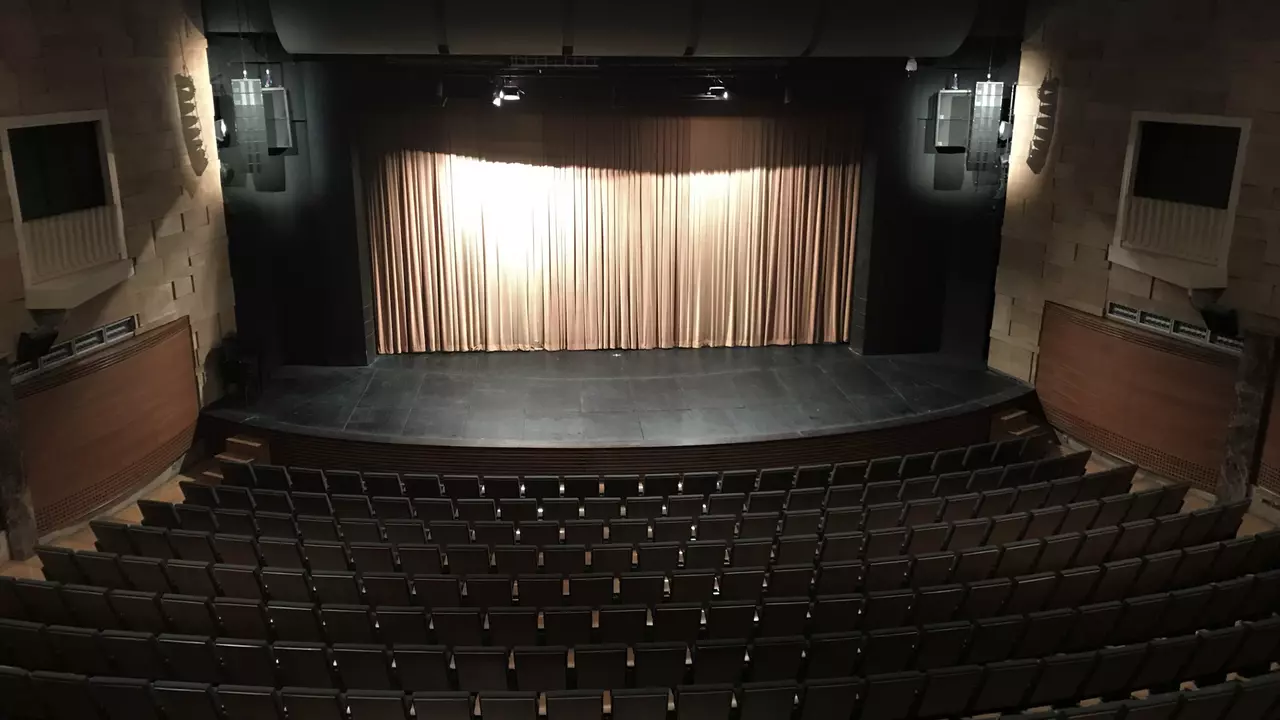 Empty theater with rows of seats facing a stage, featuring a curtain backdrop and stage lighting.