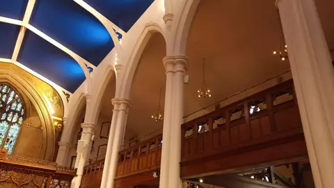 Interior view of a church featuring tall white columns, a blue ceiling, and wooden balcony seating. Stained glass windows are visible in the background.