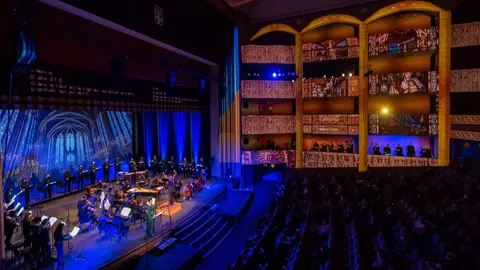 A concert performance featuring musicians on stage, with an audience seated in a theater adorned with colorful lighting and decorative architecture.