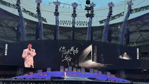 A performer in a white outfit stands on stage, with large screens displaying text and imagery in a stadium filled with an audience.