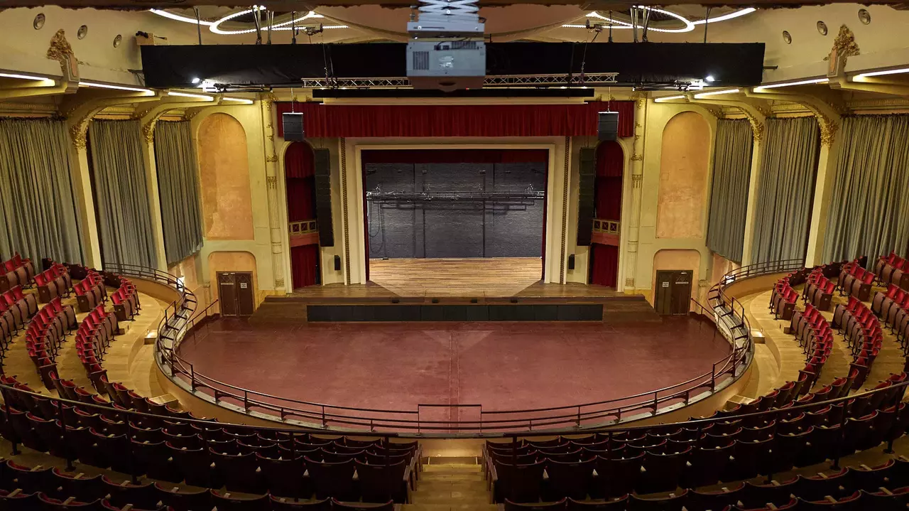 Interior view of a theater featuring a stage, red curtains, and rows of empty seats arranged in a semi-circular layout.