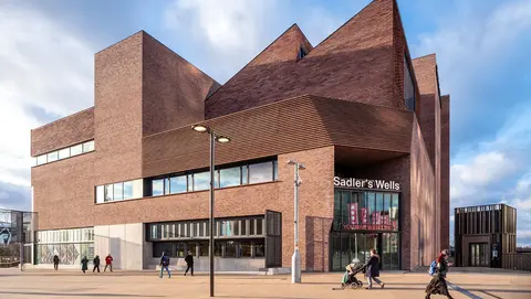 Modern brick building of Sadler's Wells theatre, featuring angular architecture and large windows, with people walking in front.