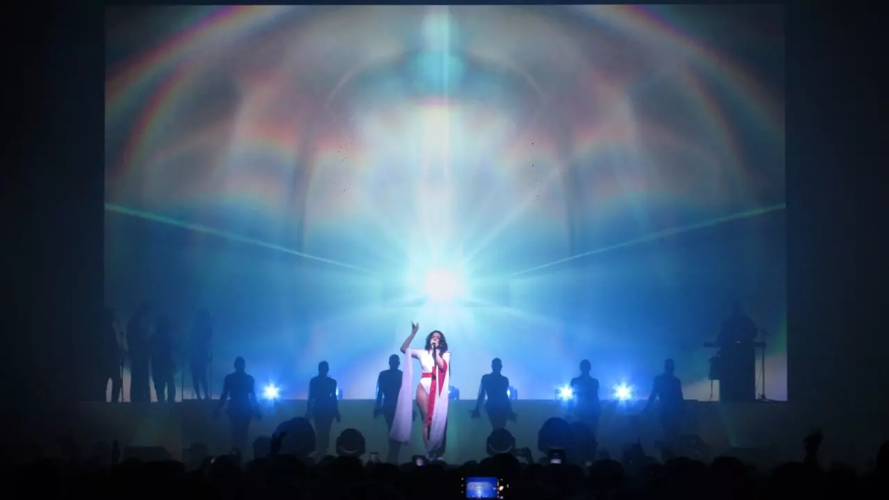 A performer stands center stage, illuminated by bright lights, with silhouetted backup dancers behind them against a colorful backdrop.