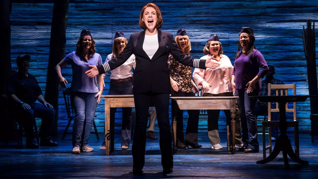 A performer in a pilot's uniform sings passionately on stage, surrounded by six supporting female cast members in casual attire.