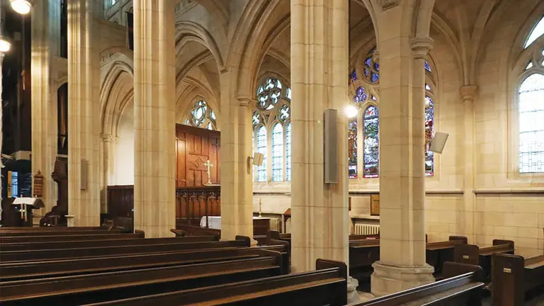 Interior of a church featuring tall stone arches, wooden pews, and stained glass windows illuminating the space.