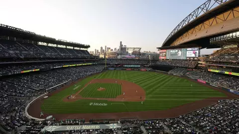 A panoramic view of T-Mobile Park during a baseball game, with a large crowd in the stands and the Seattle skyline visible in the background.