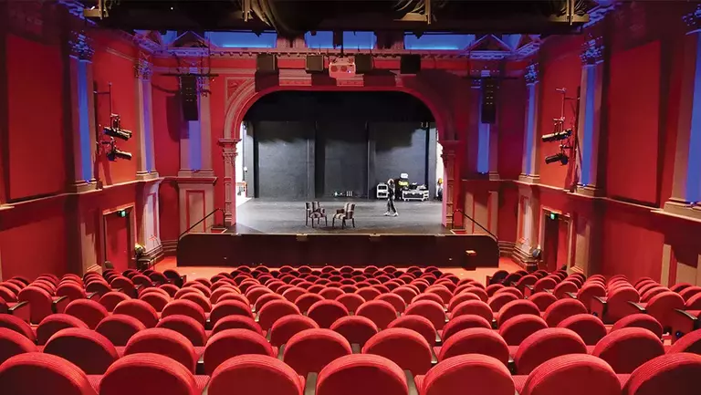 Red velvet seats fill a theater, facing a stage with a black backdrop. A few people are on stage, preparing for a performance.