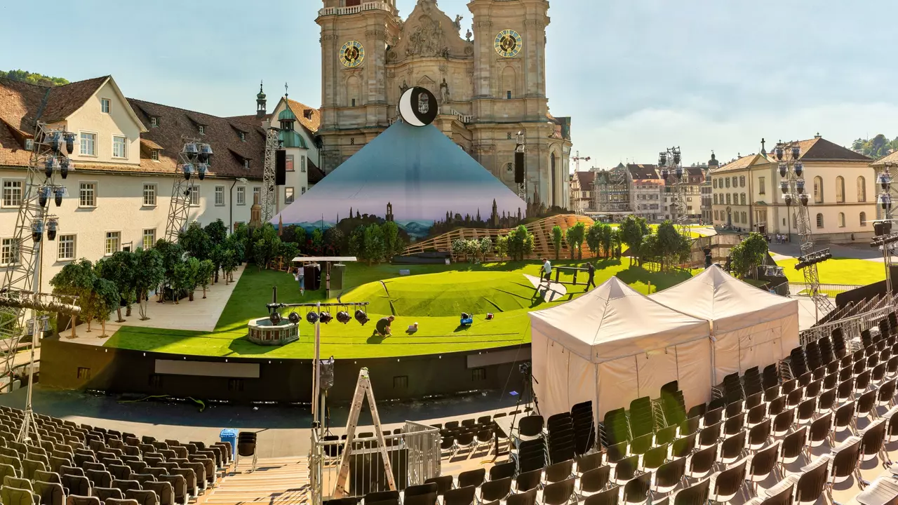 A stage set up for an outdoor event, featuring a green lawn, backdrop of a mountain, and seating arranged in front of a historic building.