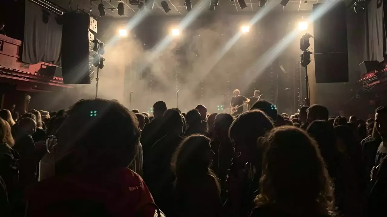 Crowd gathered in a dimly lit venue, with stage lights illuminating a smoky atmosphere, preparing for a live performance.
