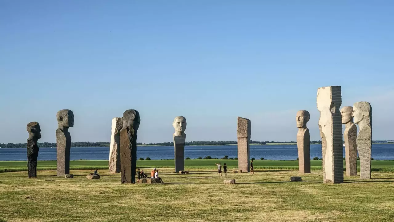 Stone sculptures resembling human heads arranged in a circular formation, with a lake visible in the background and visitors nearby.