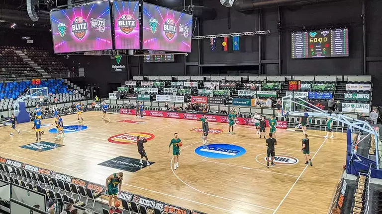 Basketball players warming up on the court, with digital scoreboards and banners visible in the arena.