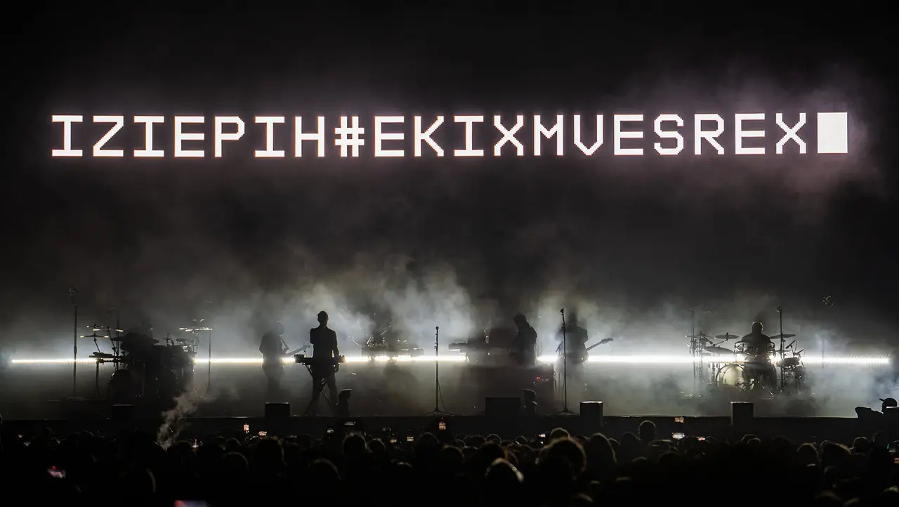 Silhouettes of musicians on stage with a large illuminated text display reading "IZIEPIH#EKIXMVESREX" against a smoky background.